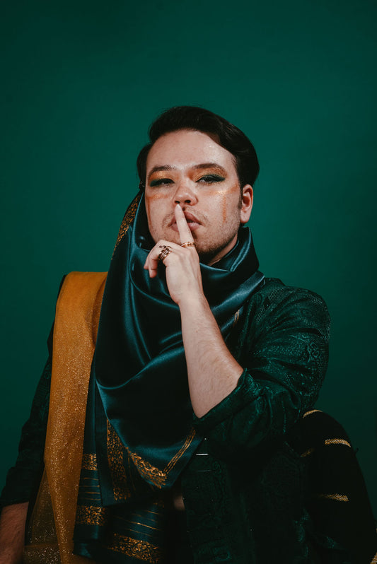 Asian man in decadent green and gold draped outfit holding finger in front of their mouth.